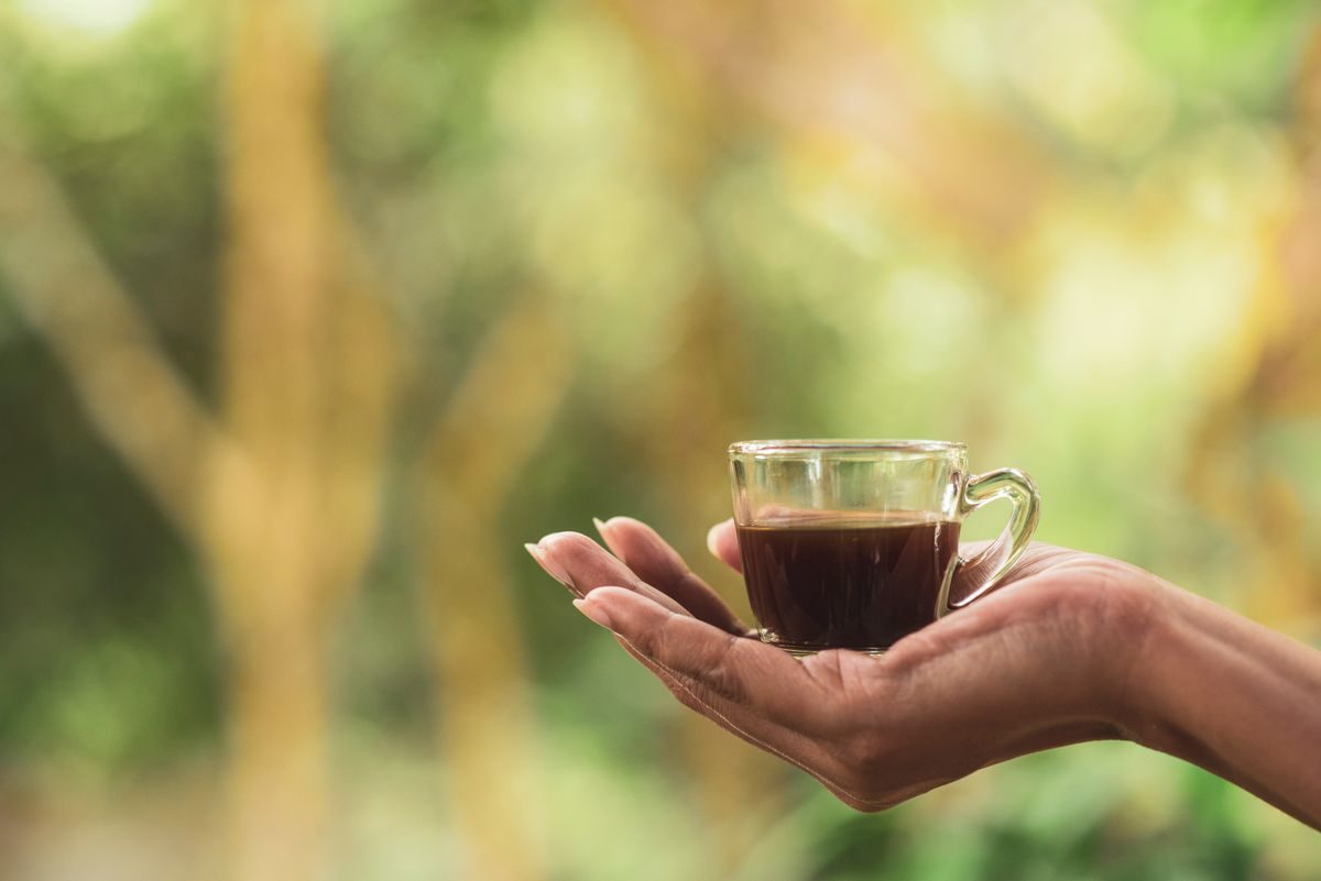 Close-up of a hand holding a glass cup of coffee with a blurred outdoor background.