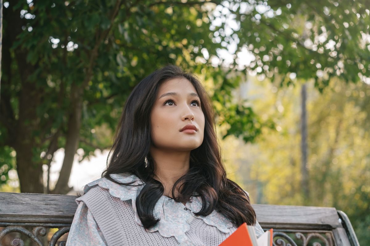 A woman sitting on a park bench, looking up contemplatively with a book in hand.