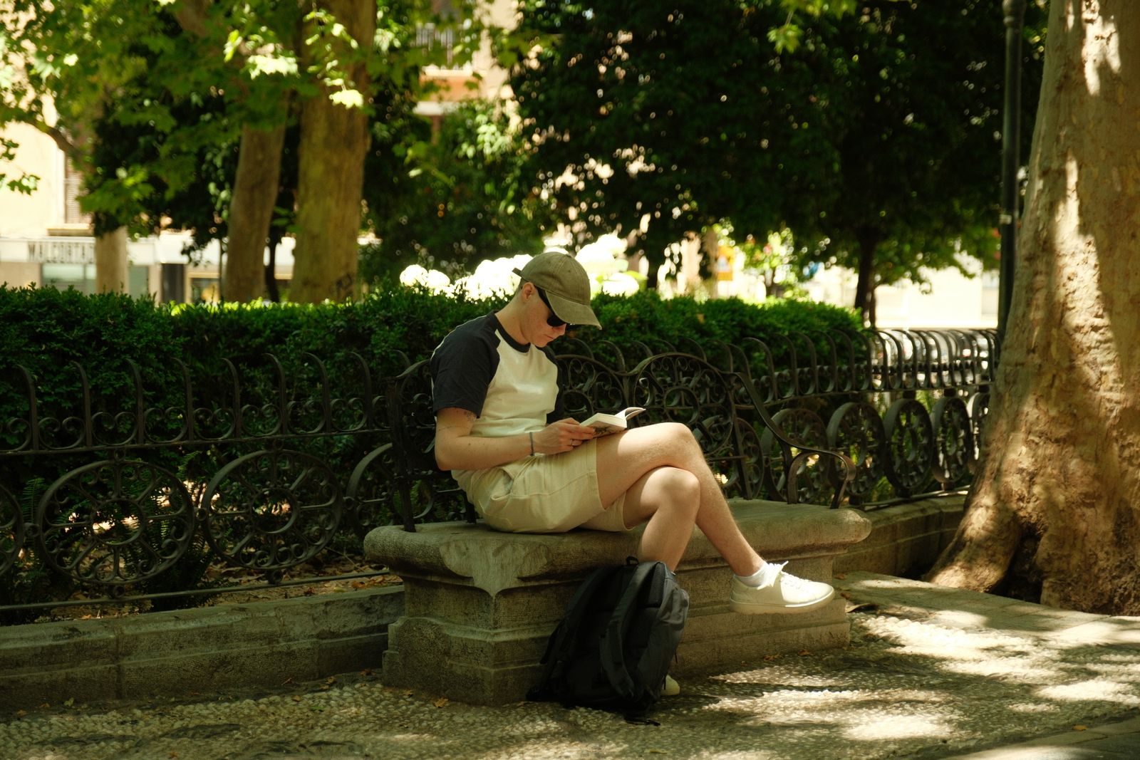 A young man enjoys reading on a bench in a Granada park, surrounded by lush greenery and sunlight.