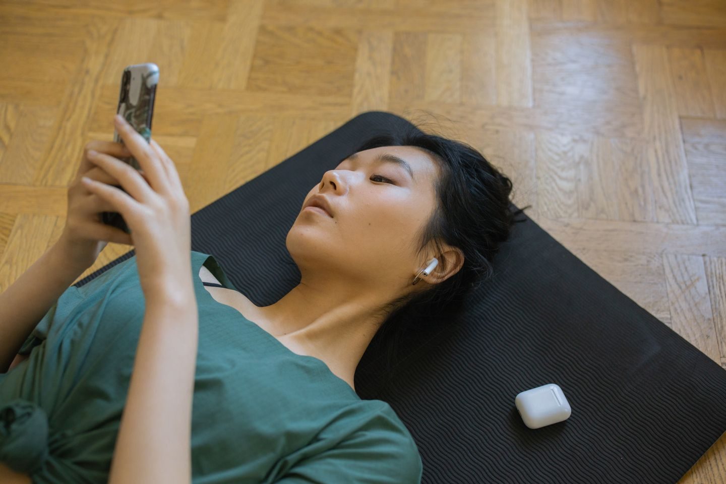A woman lying on a yoga mat using her smartphone with wireless earbuds in her ears