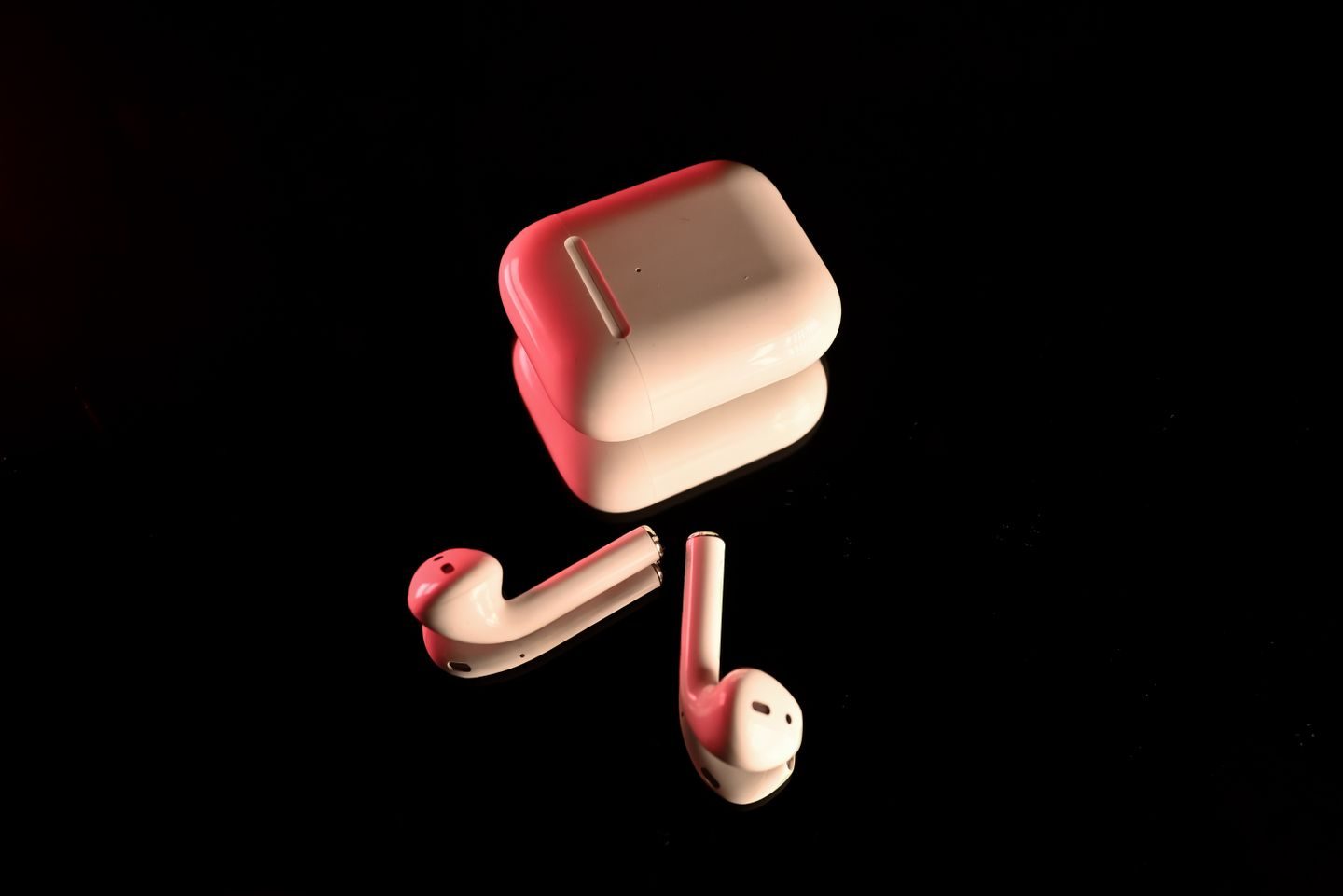 Photograph: White wireless earbuds with a charging case, highlighted with red lighting on a reflective black surface.
